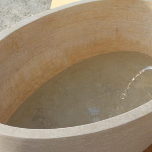 May include: An oval-shaped, beige stone bathtub filled with water. A stream of water flows from a pipe into the tub. The tub has a smooth, curved design, suggesting a spa or relaxation theme.