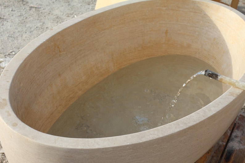 May include: An oval-shaped, beige stone bathtub filled with water. A stream of water flows from a pipe into the tub. The tub has a smooth, curved design, suggesting a spa or relaxation theme.