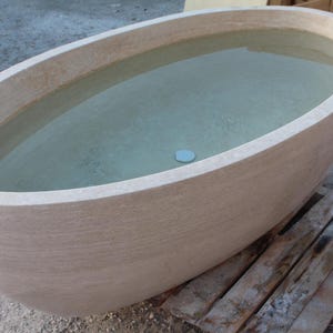 May include: An oval-shaped, beige stone bathtub filled with clear water. The tub has a smooth, rounded rim and a textured exterior. A small, round, light blue drain cover is visible at the bottom of the tub.