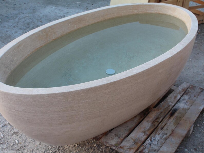 May include: An oval-shaped, beige stone bathtub filled with clear water. The tub has a smooth, rounded rim and a textured exterior. A small, round, light blue drain cover is visible at the bottom of the tub.