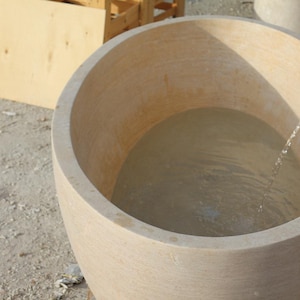 May include: A large, light-coloured, round stone tub filled with water. A stream of water flows into the tub from a pipe. The tub is set outdoors on a light-coloured surface, with wooden structures visible in the background.