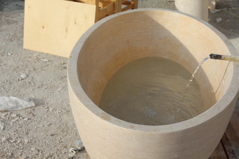 May include: A large, light-coloured, round stone tub filled with water. A stream of water flows into the tub from a pipe. The tub is set outdoors on a light-coloured surface, with wooden structures visible in the background.