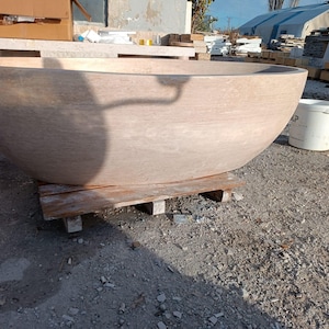 May include: A large, light brown stone bathtub with a rounded, bowl-like shape. The tub sits on a wooden pallet outdoors. The stone has a natural, textured appearance. A white bucket is visible nearby.