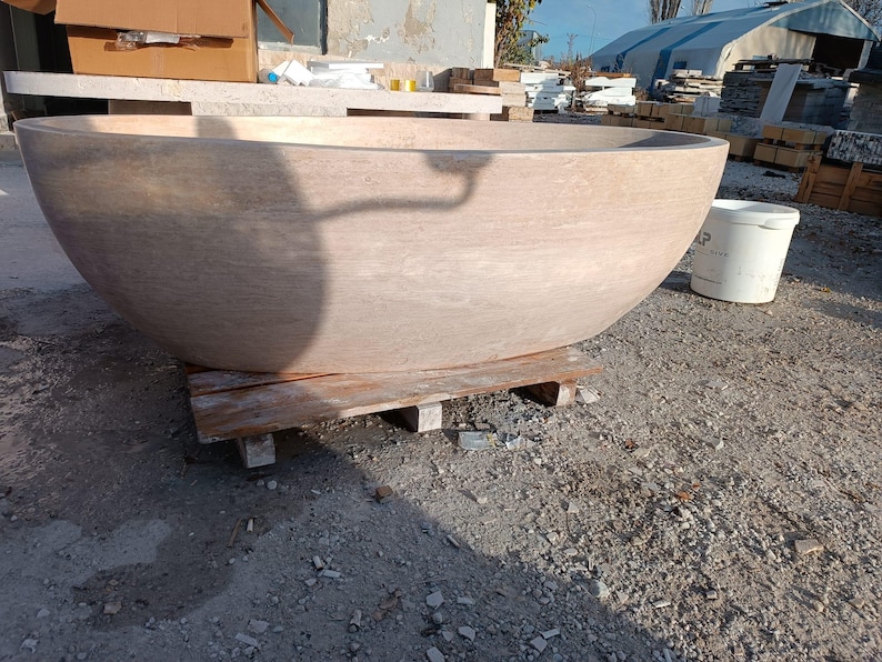 May include: A large, light brown stone bathtub with a rounded, bowl-like shape. The tub sits on a wooden pallet outdoors. The stone has a natural, textured appearance. A white bucket is visible nearby.