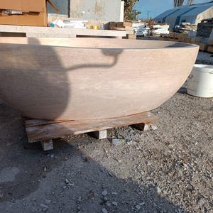 May include: A large, light brown stone bathtub with a rounded shape, resting on a wooden pallet. The tub has a smooth surface and is outdoors, with a white bucket nearby.