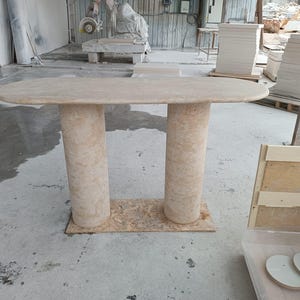 May include: A beige stone table with an oval top and two cylindrical legs. The table is set on a rectangular wooden base. The background shows a workshop with other stone pieces and tools.