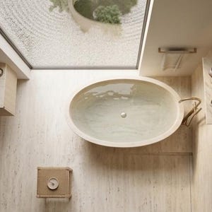 May include: An overhead view of a modern bathroom with an oval-shaped bathtub filled with water. The bathroom features a light colour palette, a skylight, and a woven stool with a clock. A sink and a towel rail are also visible.