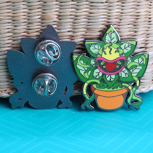 May include: A green, yellow, and red enamel pin of a carnivorous plant with a toothy mouth, in a brown pot. The back of the pin has two metal clasps.