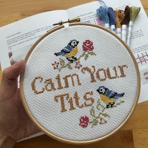 May include: A round wooden embroidery hoop containing white Aida fabric with a cross-stitch design. The design features two blue and yellow birds, small red flowers, and the brown stitched text "Calm Your Tits" centred on the fabric.