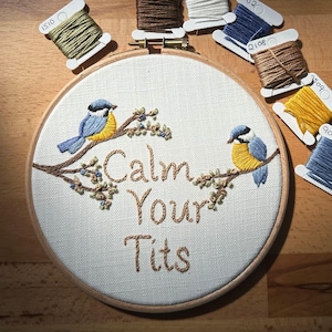 May include: Embroidered hoop art featuring two blue and yellow birds perched on brown branches with green leaves. The text "Calm Your Tits" is stitched in brown thread. Several spools of thread in various colors surround the hoop.