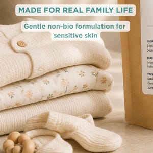 K&ouml;nnte beinhalten: Ein Stapel cremefarbener, gerippter Babykleidung, ein Kleidungsst&uuml;ck mit Blumenmuster und ein Bei&szlig;ring aus Holzperlen. Text&uuml;berlagerungen: MADE FOR REAL FAMILY LIFE und Gentle non-bio formulation for sensitive skin. Ein brauner Papierbeutel ist sichtbar.