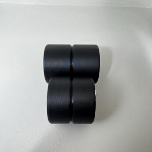 May include: Four black cylindrical objects are stacked in a rectangular formation. The objects have a smooth, matte finish and appear to be made of a solid material. The objects are arranged against a white background.