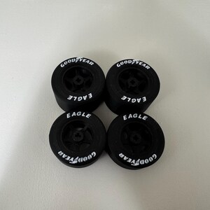 May include: Four black Goodyear Eagle racing tires. Each tire has the Goodyear logo in white on the sidewall, along with the word "EAGLE". The tires are arranged in a square formation against a white background.