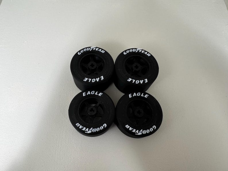May include: Four black Goodyear Eagle racing tires. Each tire has the Goodyear logo in white on the sidewall, along with the word "EAGLE". The tires are arranged in a square formation against a white background.