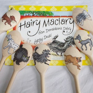 May include: A collection of wooden spoons, each adorned with a hand-painted dog design, surrounding a book. The book is titled "Hairy Maclary from Donaldson's Dairy" by Lynley Dodd. The spoons feature various dog breeds.