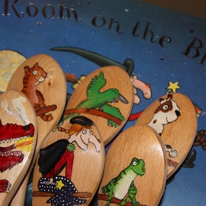 May include: A collection of wooden spoons, each hand-painted with characters from the children's book "Room on the Broom." The spoons depict a dragon, witch, cat, dog, and other characters. The book's cover is visible in the background.