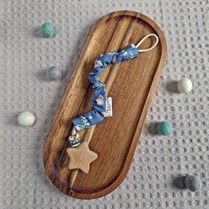 Könnte beinhalten: Ein Schnullerhalter mit einem sternförmigen Holzanhänger und einem blauen Stoffband mit einem Waldtierdruck. Der Clip liegt auf einem ovalen Holztablett. Kleine Filzkugeln in verschiedenen Farben sind verstreut.