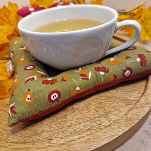 Könnte beinhalten: Eine weiße Teetasse mit Tee steht auf einem grünen, floral gemusterten Untersetzer mit rotem Rand. Der Untersetzer zeigt ein Design aus Äpfeln, Birnen und Blumen. Die Szene befindet sich auf einem Holztablett und erzeugt eine herbstliche Atmosphäre.