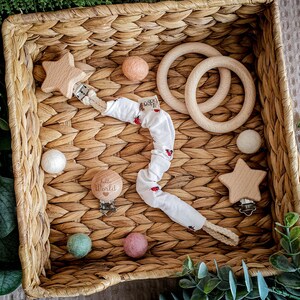 Könnte beinhalten: Ein Set Baby-Beißringe in einem Weidenkorb. Das Set beinhaltet einen Holz-Stern-Beißring, einen Holz-Ring-Beißring, einen Holz-Clip-Beißring mit der Gravur "Hello World" und einen Stoff-Beißring mit Marienkäfer-Druck.