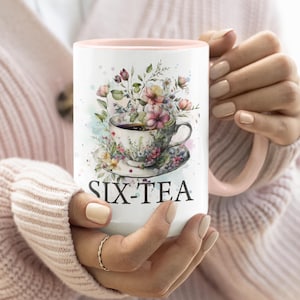 May include: White ceramic mug with a pink interior and handle, featuring a floral teacup design and the text "SIX-TEA". The teacup is adorned with colorful flowers and sits on a matching saucer. The mug is held by a person.