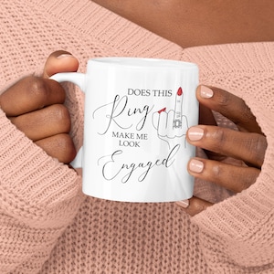 May include: White ceramic mug with the text "Does This Ring Make Me Look Engaged" in a script font. The mug features a graphic of a hand with a ring on the ring finger. The nails are painted red. The mug is held by a person wearing a pink jumper.