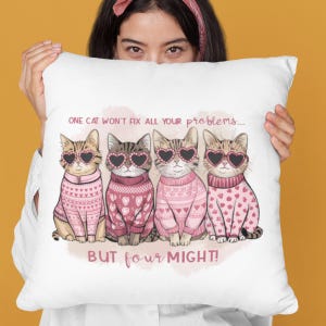 May include: White throw pillow with a pink and white illustration of four cats wearing pink sweaters and heart-shaped sunglasses. The text on the pillow reads "One cat won't fix all your problems... But four might!"