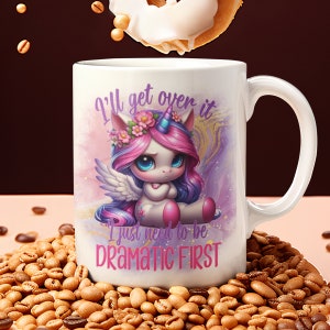 May include: White ceramic mug featuring a sad unicorn illustration with pink and purple hair, wings, and a floral crown. The mug has the text "I'll get over it, I just need to be DRAMATIC FIRST". The mug is surrounded by coffee beans.