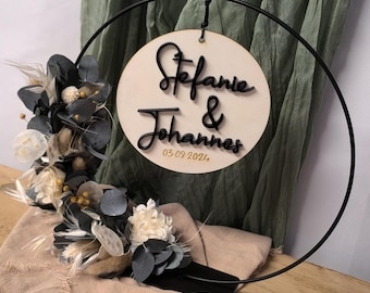 May include: A wooden circle with the text "Stefanie & Johannes 03.09.2024" in black script lettering hangs from a black metal hoop. The hoop is attached to a black stand. A bouquet of dried flowers is in the foreground.