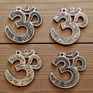 Puede incluir: Cuatro adornos con el símbolo Om con intrincados patrones en blanco y negro. Los símbolos están dispuestos sobre una superficie de madera, mostrando diseños detallados y un tema espiritual. Cada adorno tiene un patrón único y detallado.