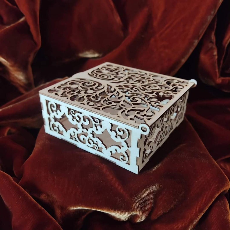 Op de afbeelding: Een witte houten doos met ingewikkelde uitgesneden ontwerpen, die lijken op een boom met takken en bladeren. De doos staat op een rood fluwelen oppervlak.