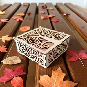May include: A square, cream-coloured wooden box featuring a tree of life design cut into the lid and sides. The box rests on a wooden surface with scattered autumn leaves. The box has a small metal loop on the top.