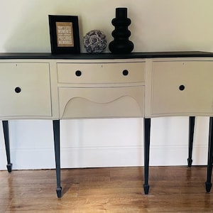May include: A cream-coloured sideboard with a black top and legs. The cabinet has two doors and a drawer with black knobs. A framed print with the text "TODAY'S LITTLE MOMENTS PRECIOUS MEMORIES" sits on top, along with a decorative sphere and a black vase.
