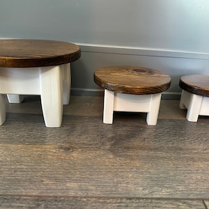 Peut inclure: Trois supports de piédestal ronds en bois avec un dessus teinté brun foncé et des pieds peints en blanc. Les supports sont de tailles différentes, le plus grand à l'arrière et le plus petit à l'avant.