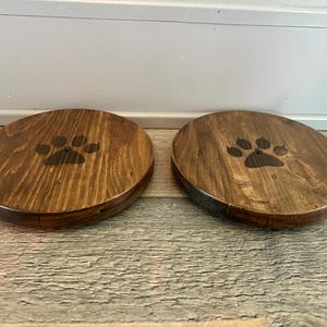 Puede incluir: Dos posavasos redondos de madera con tinte oscuro. Cada posavasos presenta un diseño de huella de pata en el centro. Los posavasos están colocados sobre una superficie de madera.