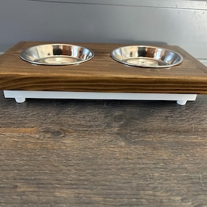 May include: A rectangular pet feeder station featuring a dark brown stained wooden top with two circular cutouts holding removable stainless steel bowls. The wooden top rests on a solid white base supported by four small, short white feet.