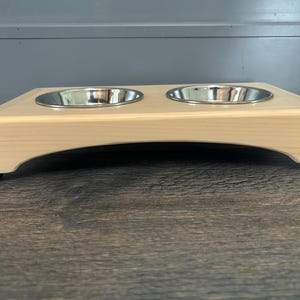 May include: A light-colored wooden pet feeder stand featuring two circular stainless steel bowls embedded into the top surface. The stand has a smooth, arched base design with small black feet at each corner for stability and elevation.