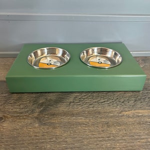 May include: A sage green rectangular elevated pet feeder stand featuring two circular stainless steel bowls. Each bowl contains a circular product label with text and a graphic. The stand has a smooth, solid-colored finish and a flat top surface.