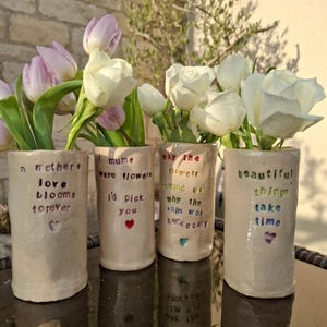 May include: Four handmade ceramic vases with floral arrangements. Each vase has a unique inscription in black and colourful accents. The vases hold white roses and pink and white tulips. The text includes phrases like "a mother's love blooms forever."