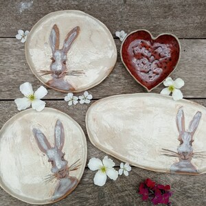 May include: Two round ceramic plates and one oval ceramic plate, each featuring a hand-painted brown and white rabbit illustration. A heart-shaped ceramic bowl with a deep red, textured glaze is included, all decorated with white flowers.