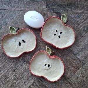 May include: Three ceramic apple-shaped dishes with red rims and cream-coloured centres, each featuring a small green leaf detail at the top and dark seed-like cutouts in the middle, accompanied by a single smooth white oval bar of soap.