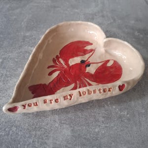 May include: A heart-shaped ceramic trinket dish with a glossy cream finish. The centre features a hand-painted bright red lobster. The rim is embossed with the red text "you are my lobster" flanked by two small red heart icons on each side.