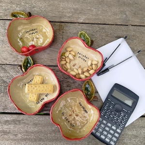 May include: Four ceramic apple-shaped bowls with red rims and green leaves. Two bowls are inscribed with "teacher treats" and two with "awesome to the core." The bowls contain cherries, pistachios, and crackers, accompanied by two pens and a calculator.