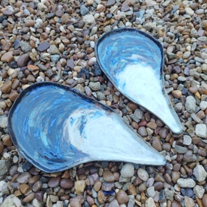 May include: Two ceramic serving dishes shaped like seashells. The dishes are white with blue and brown marbling. They are sitting on a bed of small grey and brown stones.