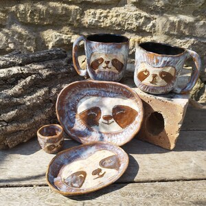 May include: Set of four ceramic mugs and plates featuring a sloth design. The mugs and plates are glazed in a light blue and brown colour scheme. The sloth faces are painted in brown with white eyes and a smile.