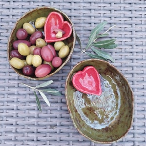 May include: Two oval ceramic bowls with a mottled olive green glaze. One bowl is filled with green and purple olives and contains a small red heart-shaped dish. The other bowl features a red heart-shaped dish and the embossed text follow your heart.