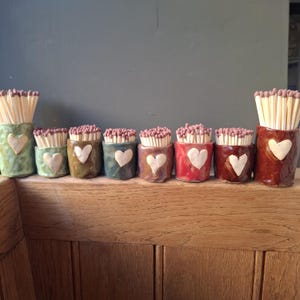May include: A collection of eight small, cylindrical ceramic match holders arranged in a row. Each holder features a textured surface, a unique glaze colour ranging from green to red and brown, and a small, raised white heart on the front.