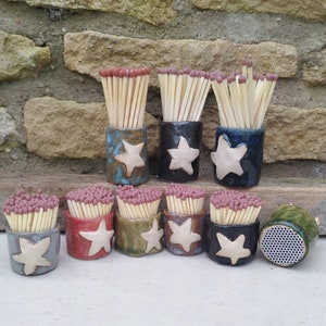 May include: Nine ceramic matchstick holders with a white star design. Each holder is filled with wooden matchsticks with red tips. The holders are arranged in a row on a wooden surface.