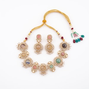 May include: A gold-toned necklace and earring set featuring round and square cabochon stones in various pastel colors, surrounded by small white crystals and pearls. The necklace has an adjustable gold cord with decorative beaded tassels at the ends.