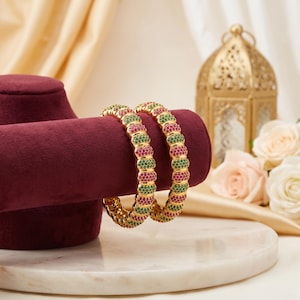 May include: Two gold bangles featuring a repeating pattern of small, round, vibrant pink and green stones. The circular bracelets have a textured, segmented design with alternating rows of pink and green gems set into a polished gold metal frame.