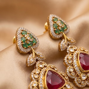 May include: A pair of gold drop earrings featuring a heart-shaped top cluster of small green emeralds surrounded by clear diamonds. Below this, a larger teardrop-shaped red ruby is set in a gold frame encrusted with numerous small clear diamonds.